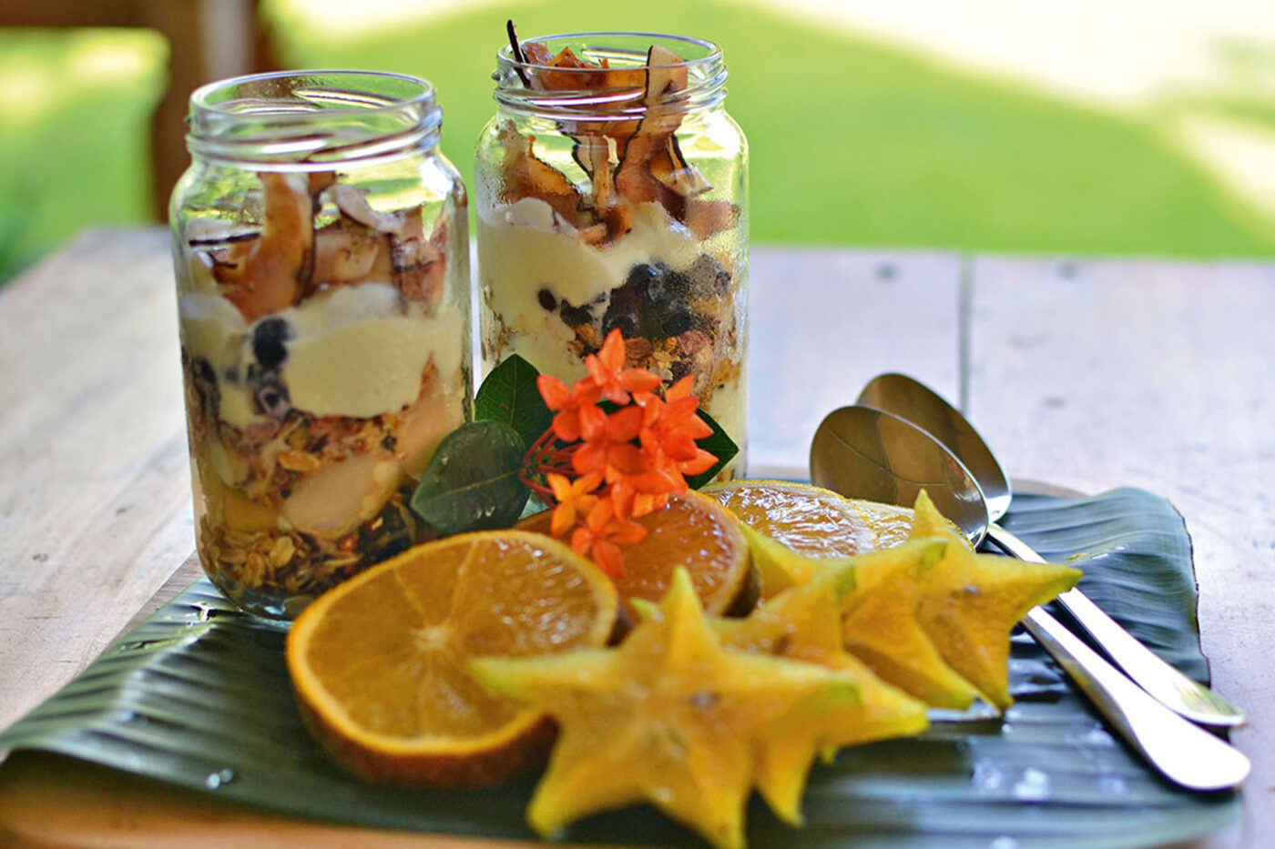 Two jars of museli and fruit sitting on a leaf placemat. Four lemons, two spoons and fruit cut out in the shape of stars are resting in front of them.