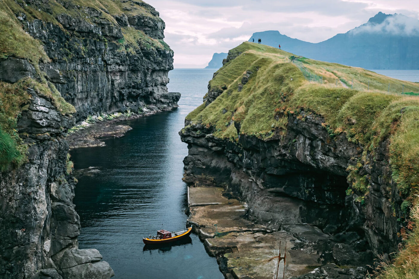 A boat in the middle of a large canal that leads into the ocean. On the right, there's four people stood on top of one of the hills.