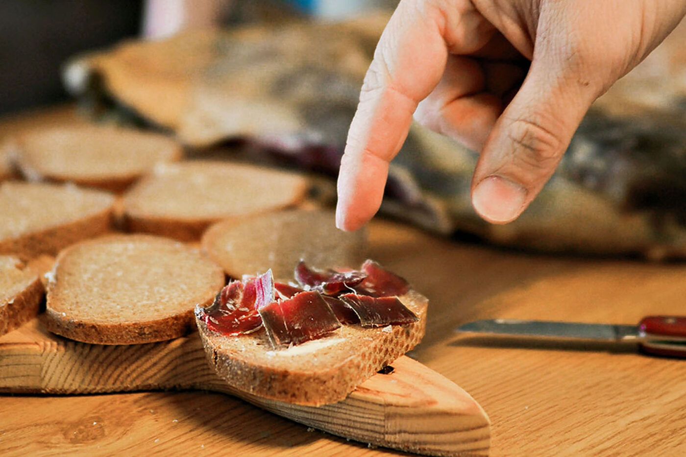 Cut pieces of bread on a chopping board. The bread in the foreground has bits of fermented lamb and butter on it. A hand is grabbing for this piece.