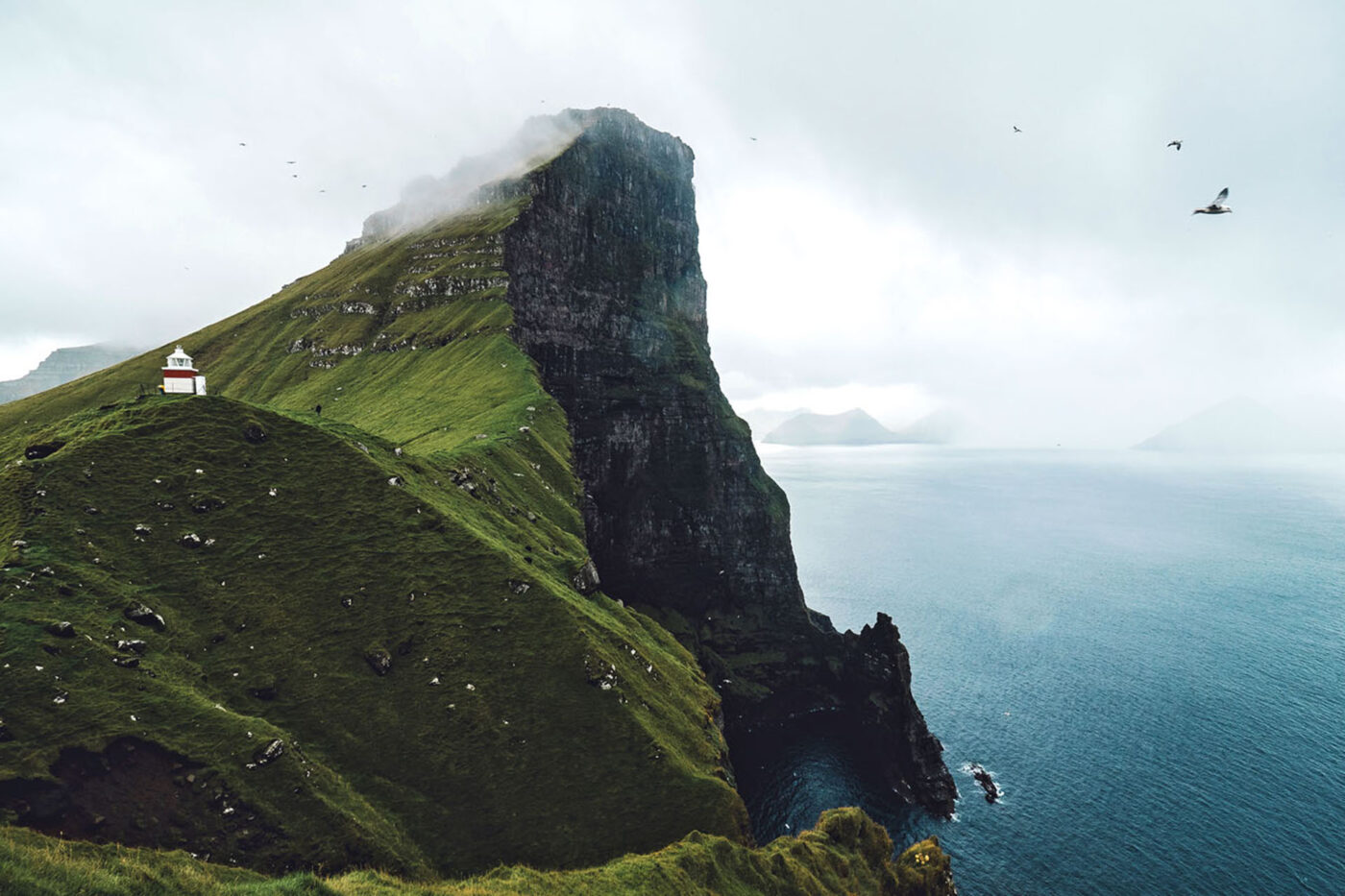 A white and burgundy lighthouse sitting on the top of a big hill. The deep blue sea is at the bottom of the hill.