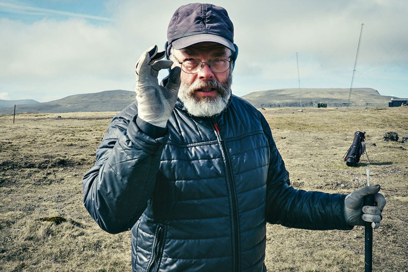 A middle-aged man with a grey beard wearing a blue hat, glasses, a blue puffer jacket and grey gloves. He's holding up his hand up to his face, making a circle with his thumb and first finger. In his other hand, he's holding a golf club.