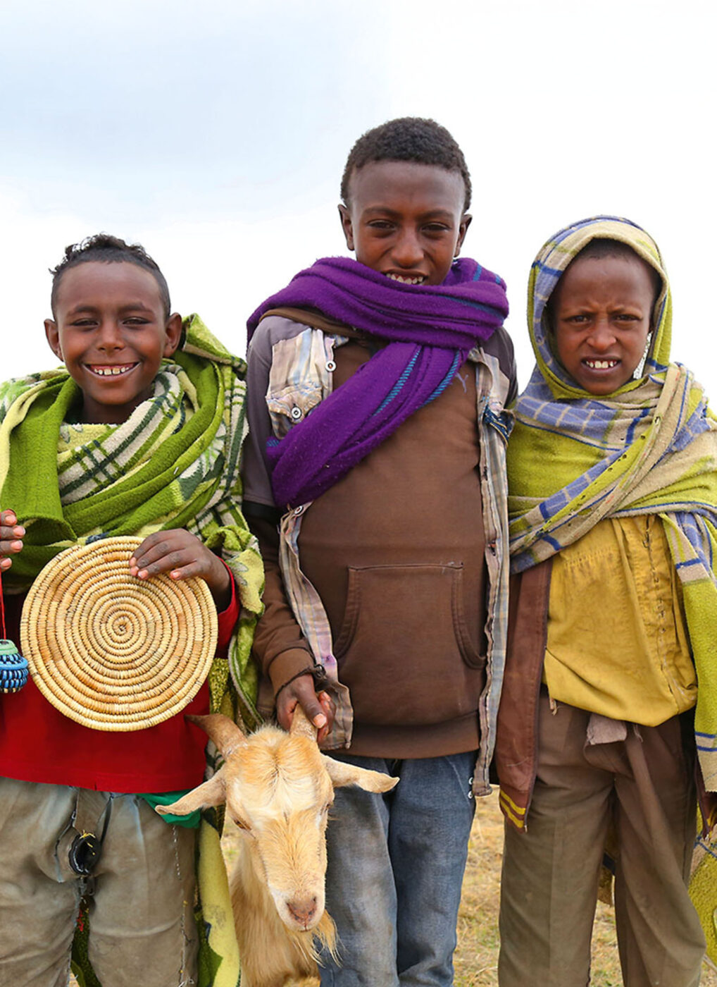 Three local children smiling for the camera. Two of them are holding a goat.