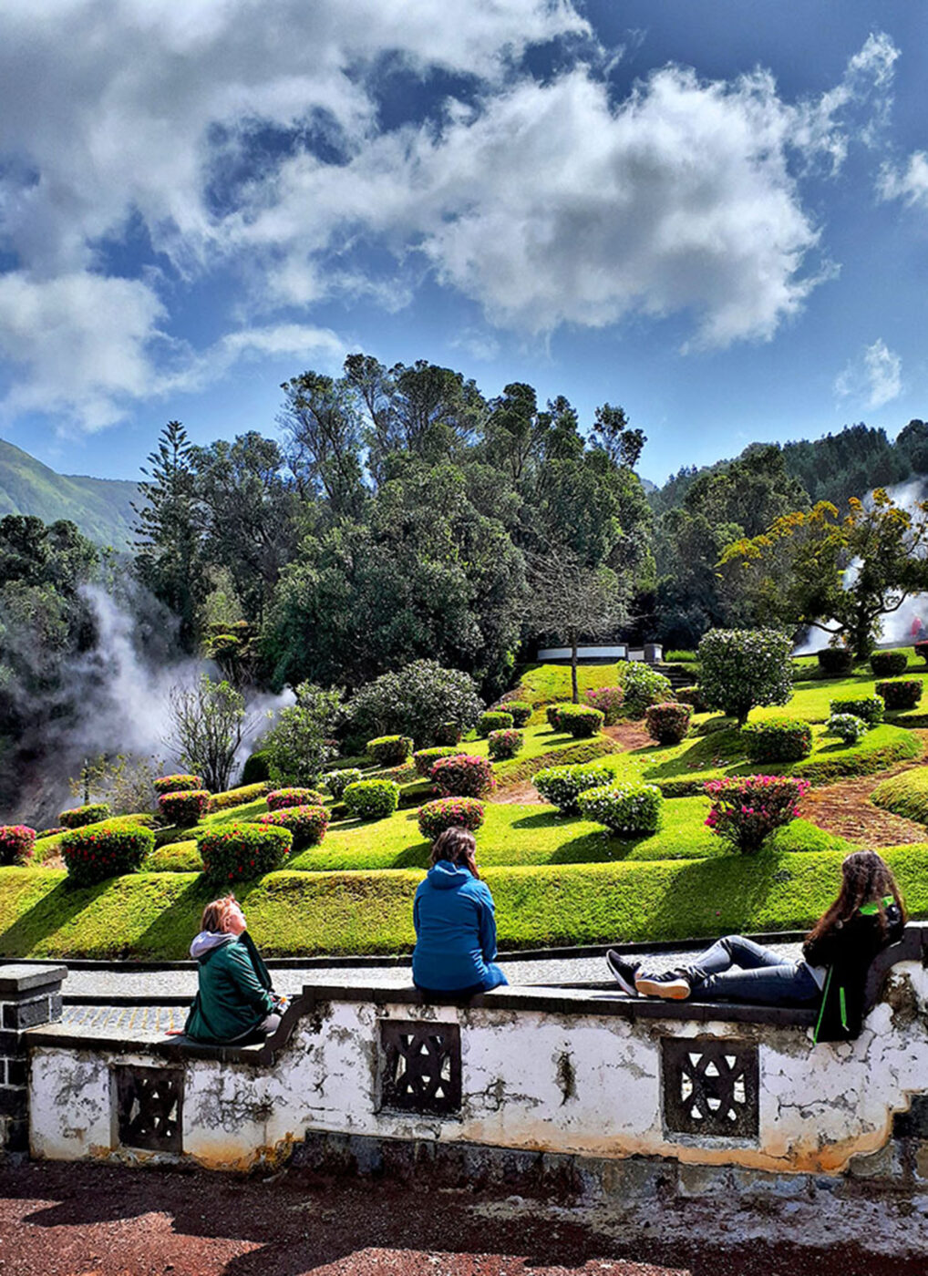 Three people sitting on a stone fence, watching the steam rise up from the ground at the hot springs.