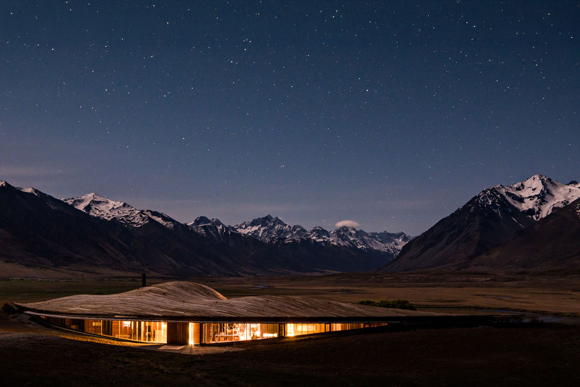 A house lit-up in front of snowy mountains at dusk.