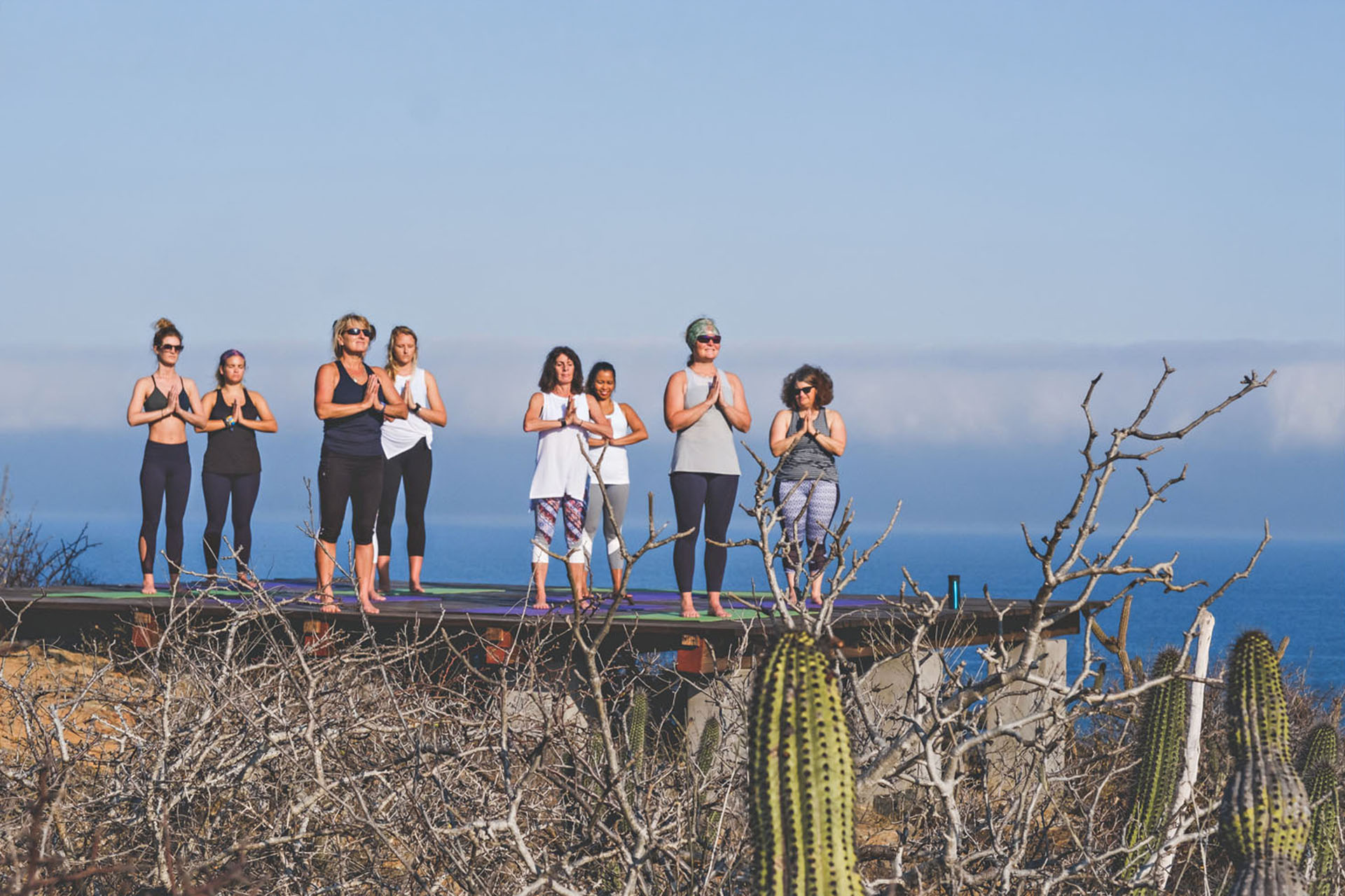A group of people holding their hands together in a praying fashion, stood on a platform by the sea.