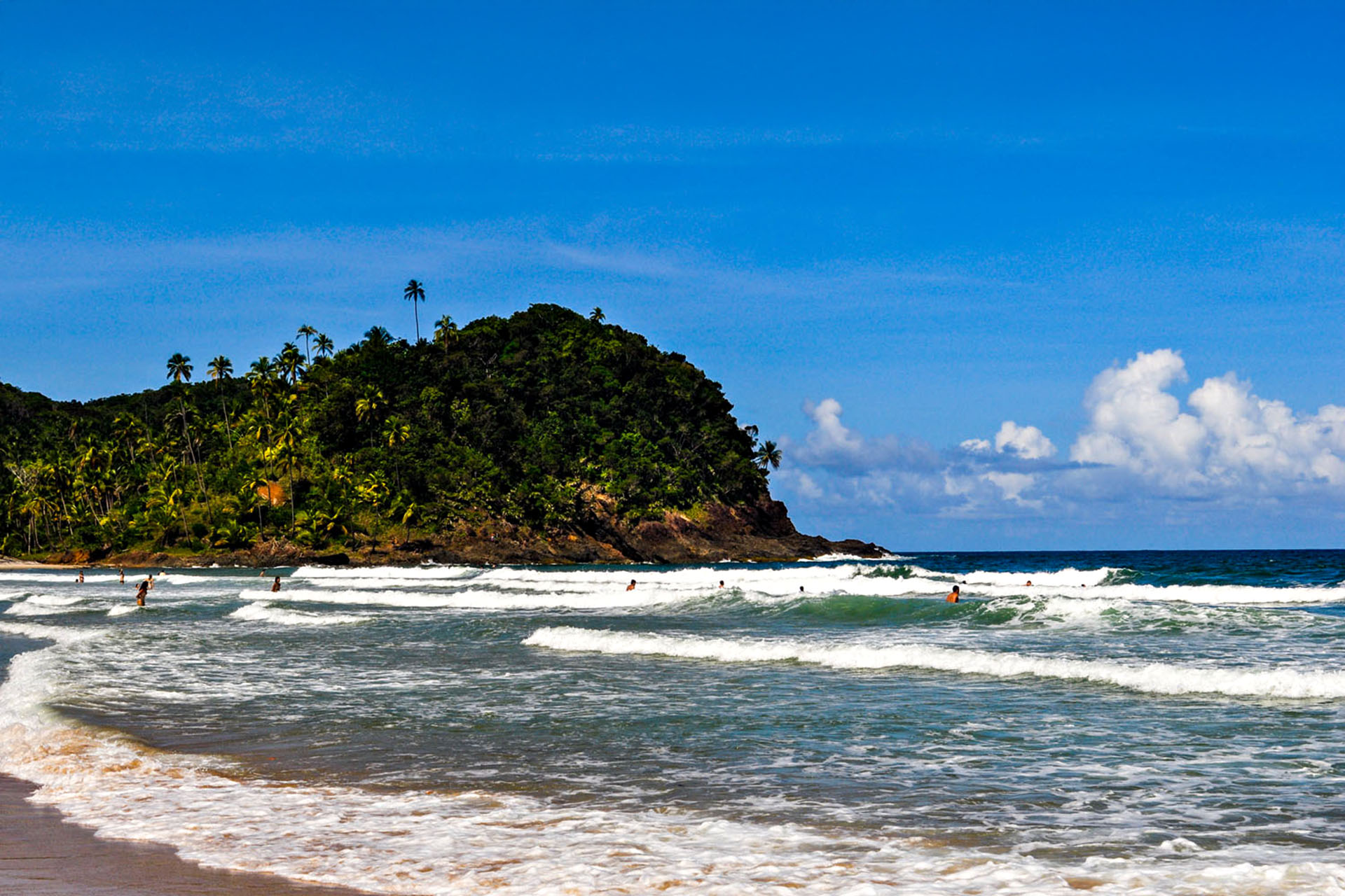 Waves coming into shore, with people playing in them. In the background, an island of palm trees.