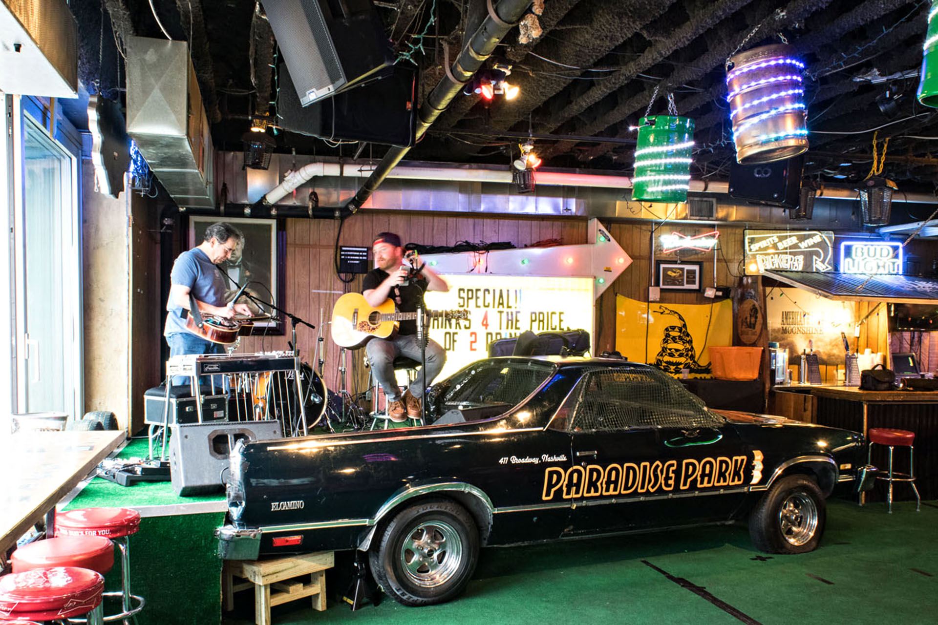 A bar filled with memorabilia and LED lights. A big car is the centerpiece. Behind it, a stage with a band about to play.