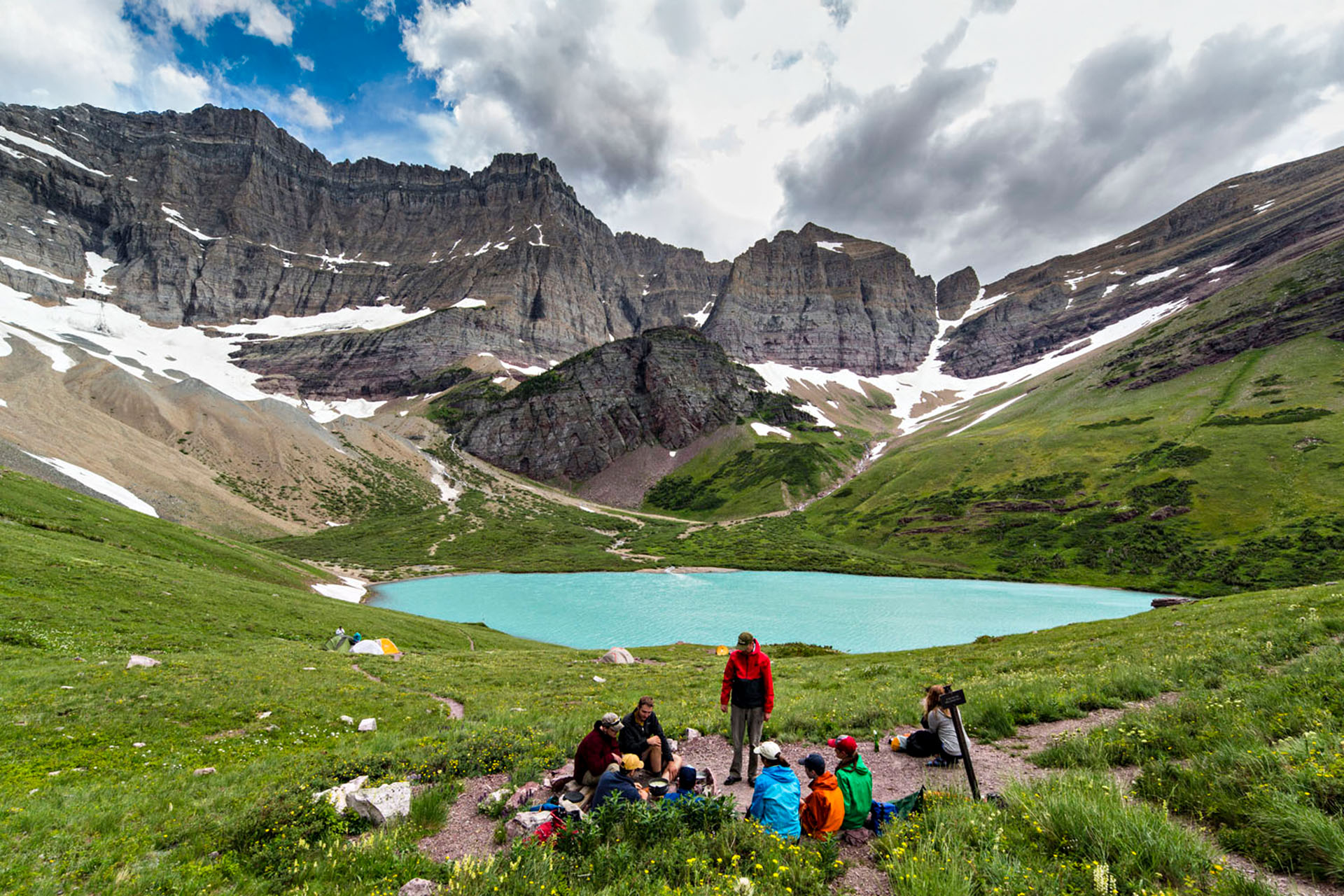 A group of people sitting on the grass. Behind them, a big lake. Behind the lake, large glaciers.
