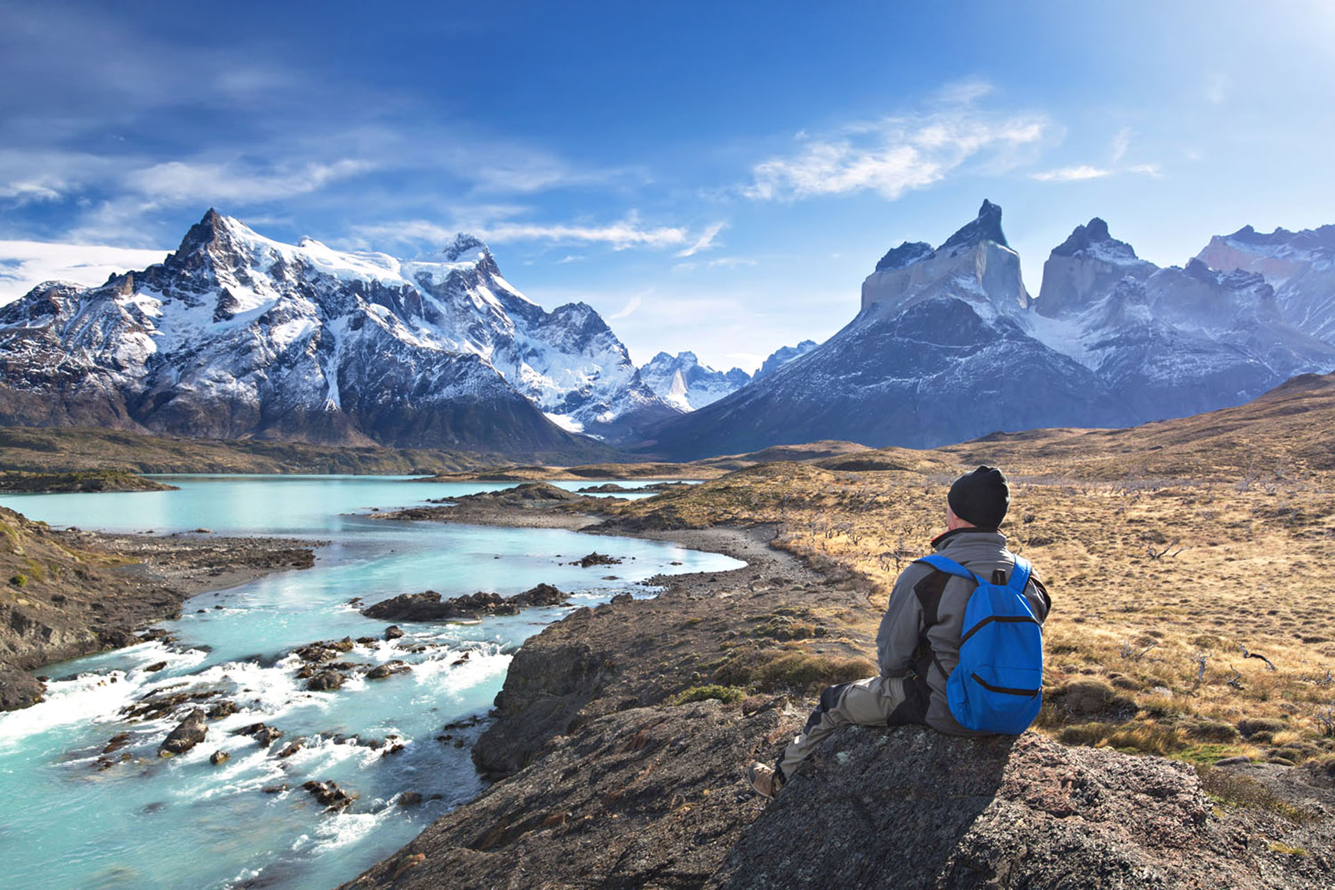 A person sitting down in front of light blue river rapids. In the background, snowy mountain ranges.