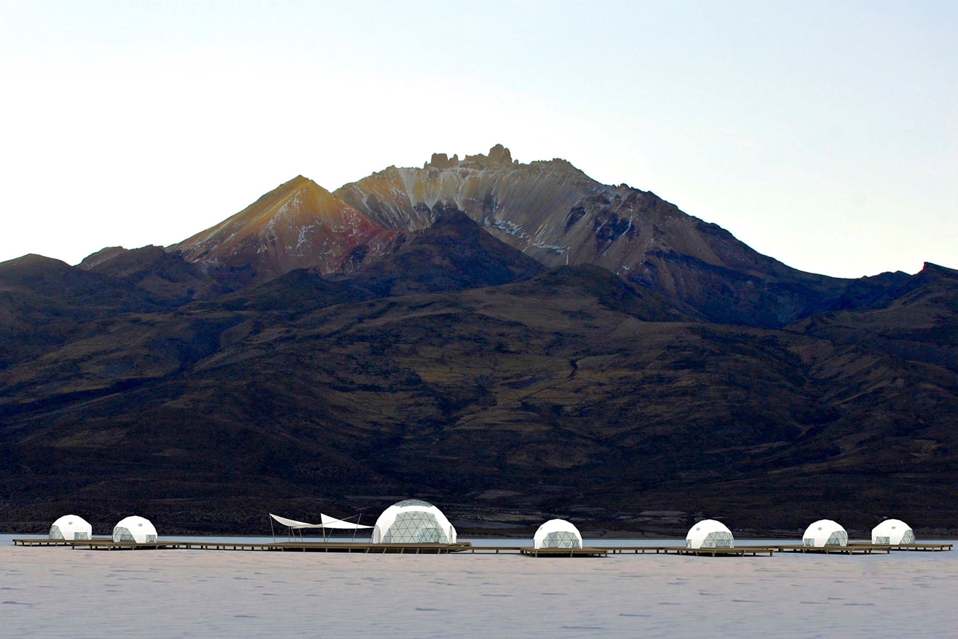 Round glamping tents that look like snowglobes, next to the water. Behind them, a huge mountain.