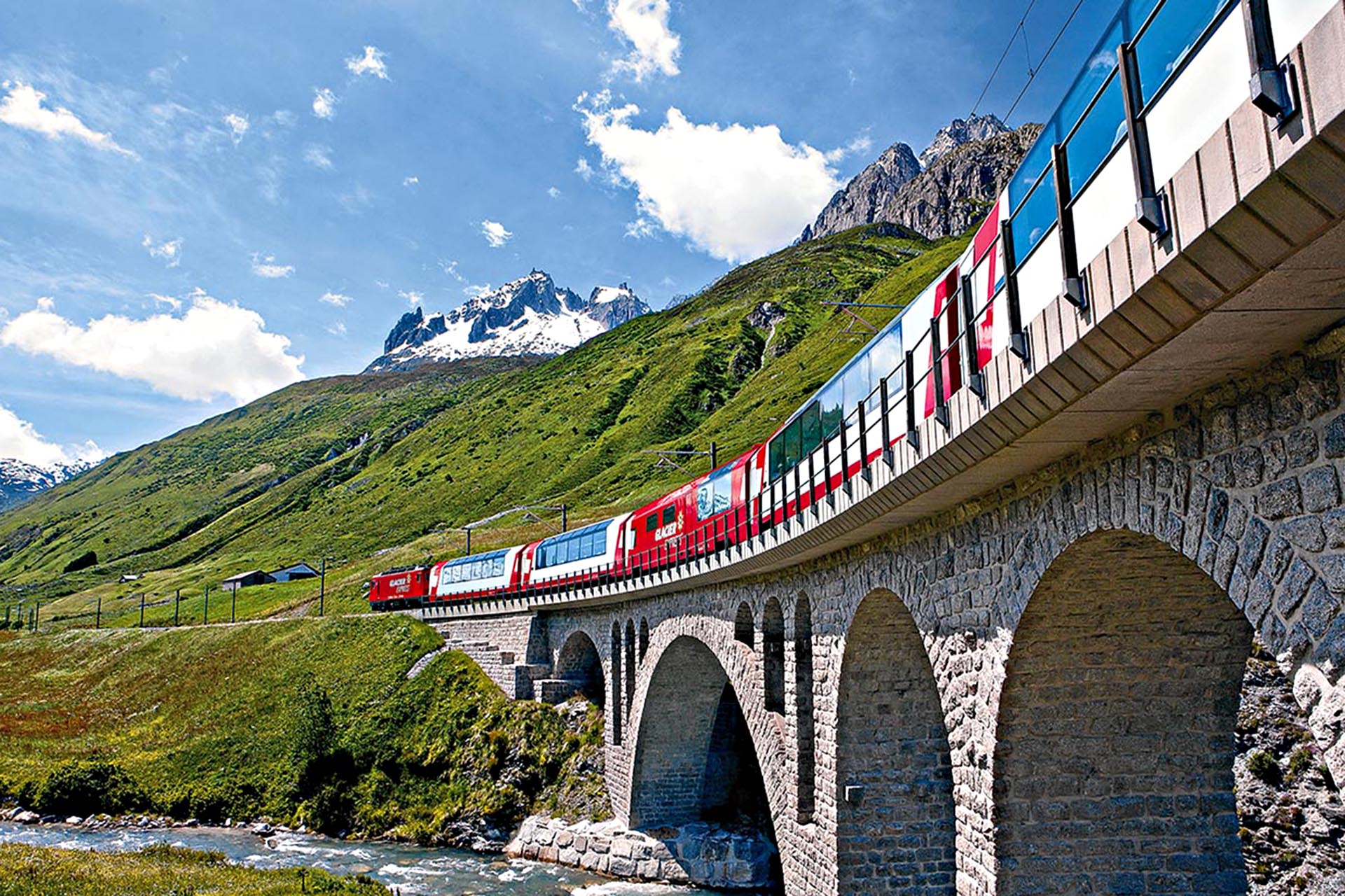 A red train crossing a bridge over a river in the mountains.