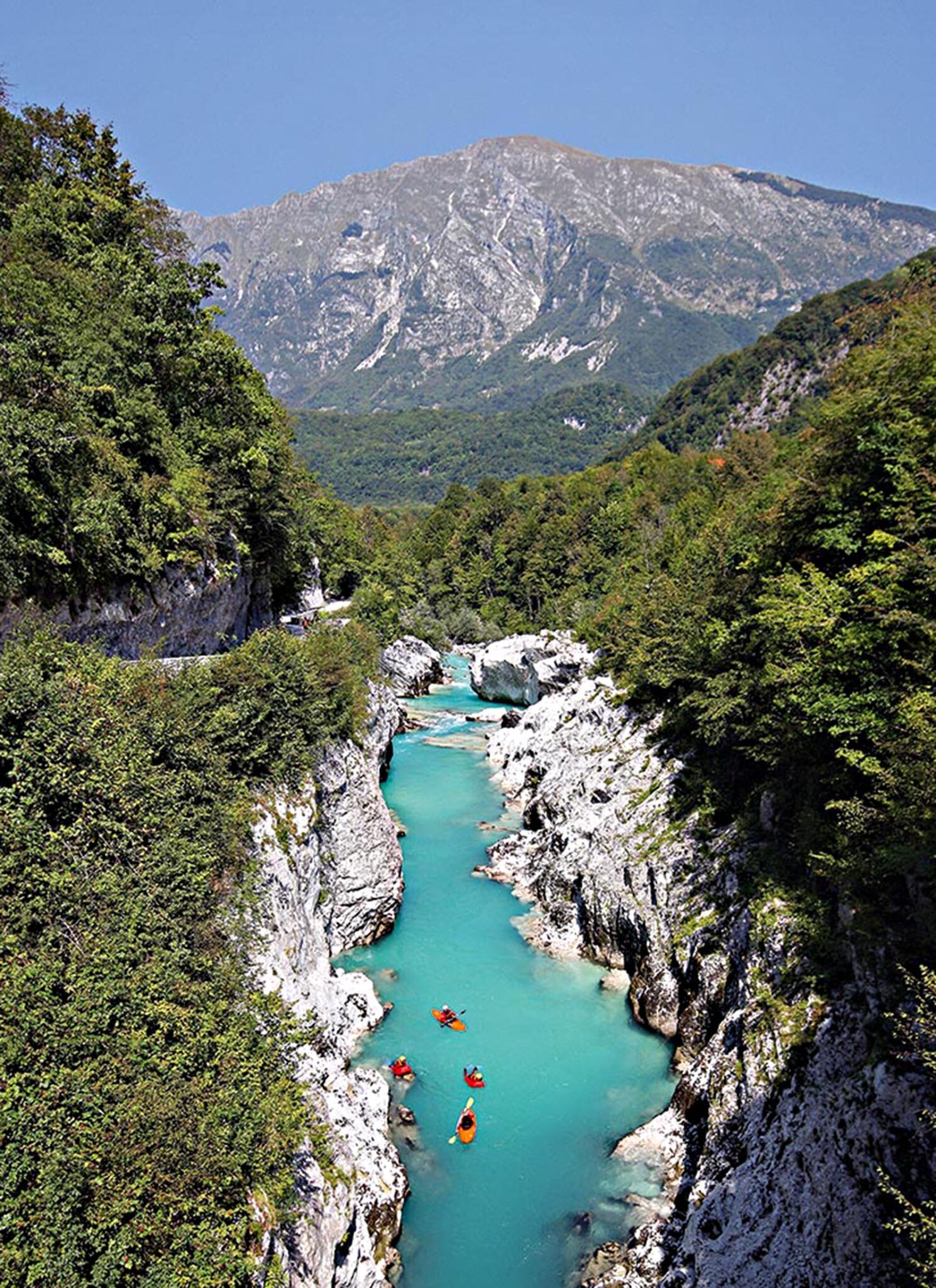 A group of people in kayaks rowing down a narrow river between rock formations. A big mountain is in the background.
