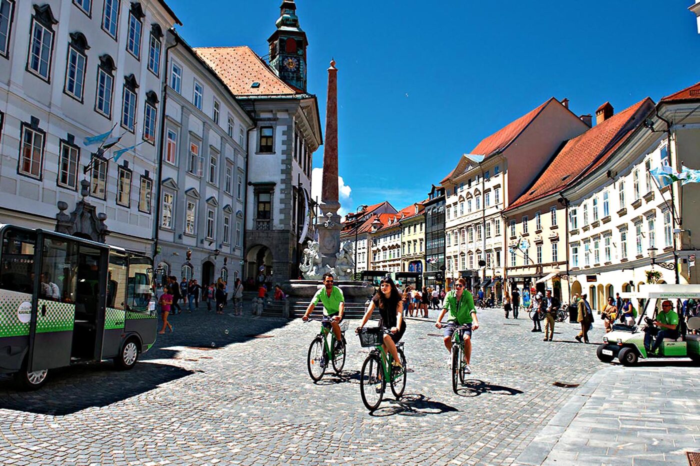 A busy street, with 3 people riding bikes in the foreground. In the background people in buggies and people on foot.