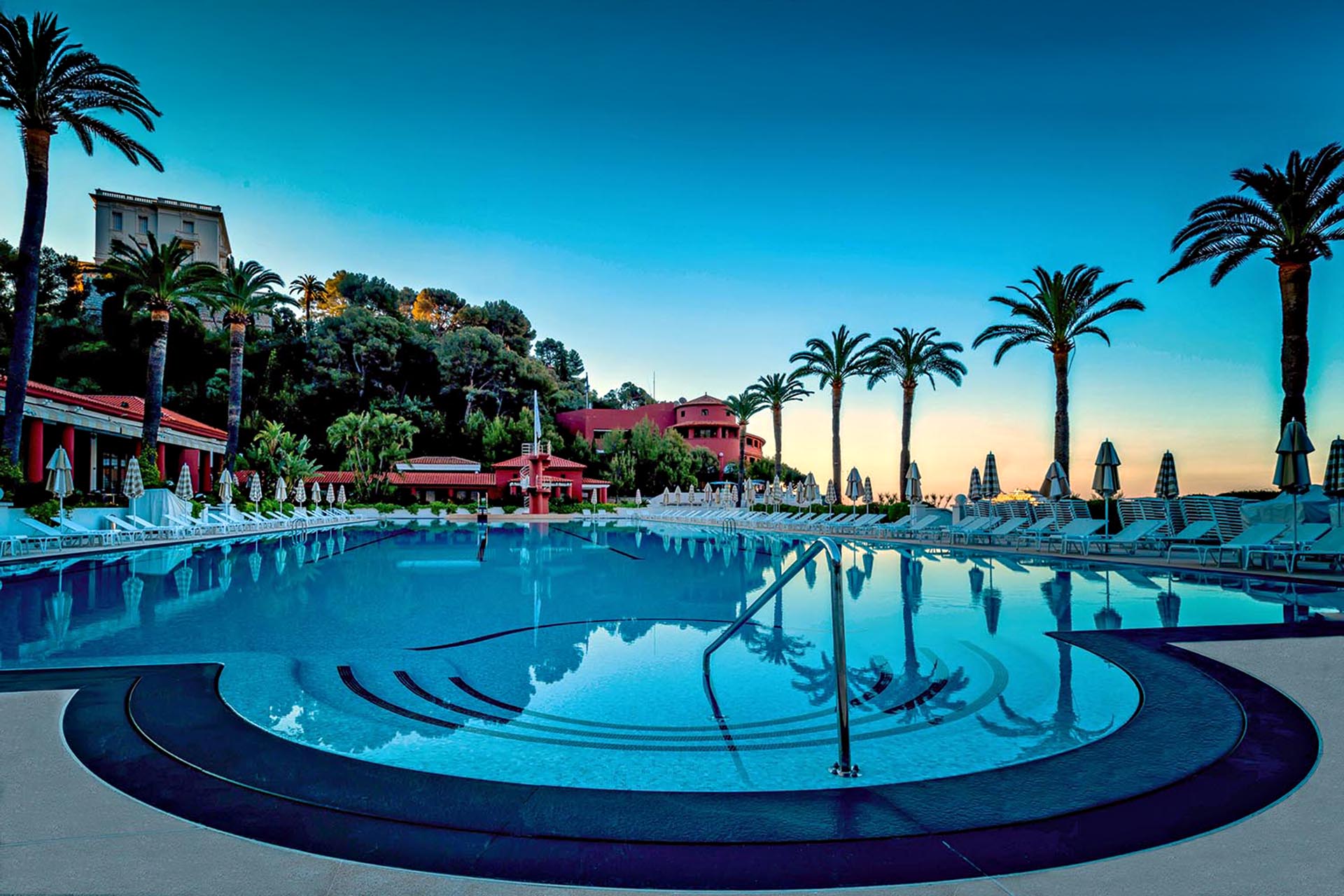 A hotel pool at sunset, with palm trees lined up either side.