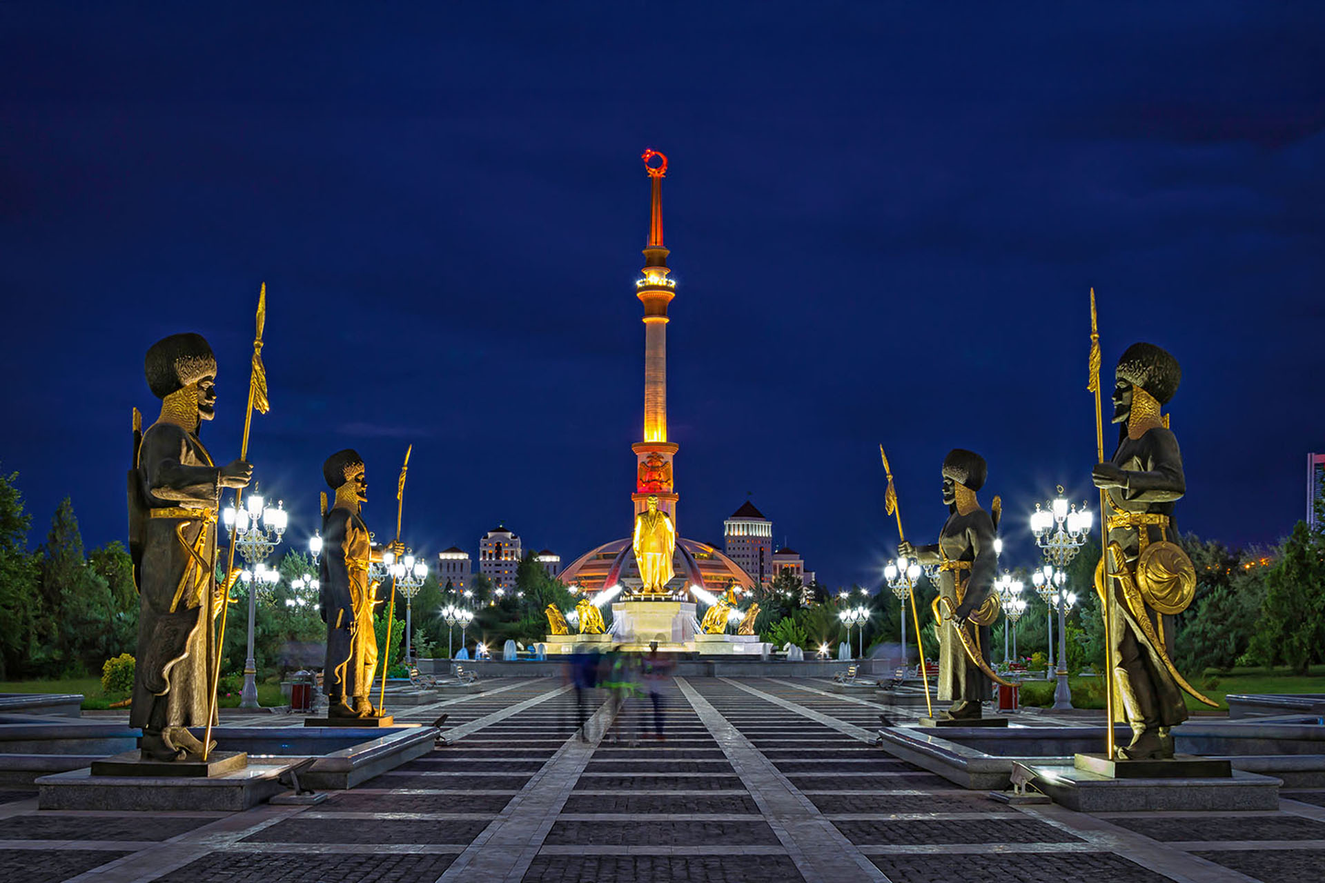 A big park with four statues facing each other. The walkway in the middle leads to a tower.