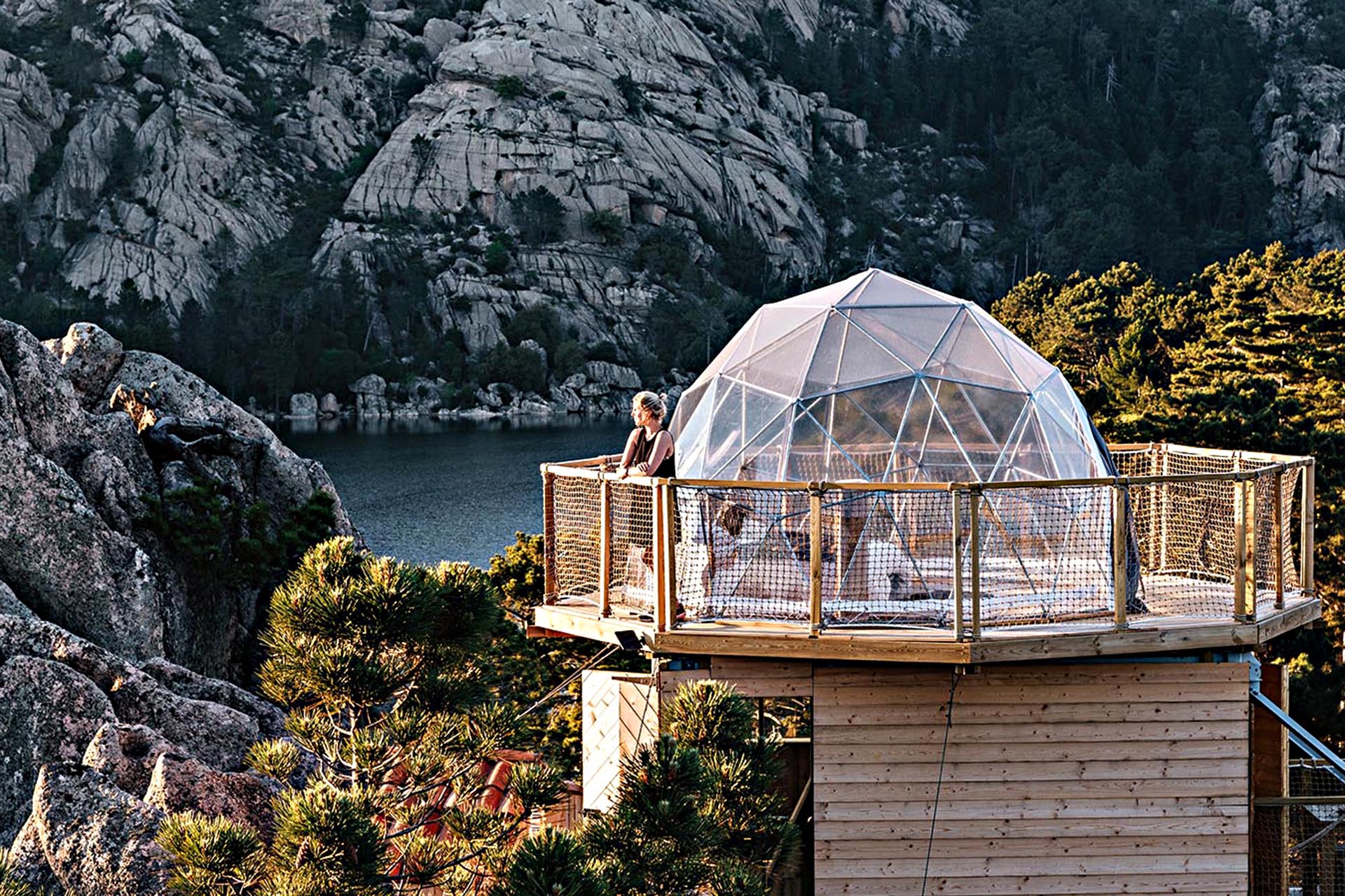 A woman on the balcony of a giant snowglobe shaped home made of glass. Rock formations and water coat the landscape around it.