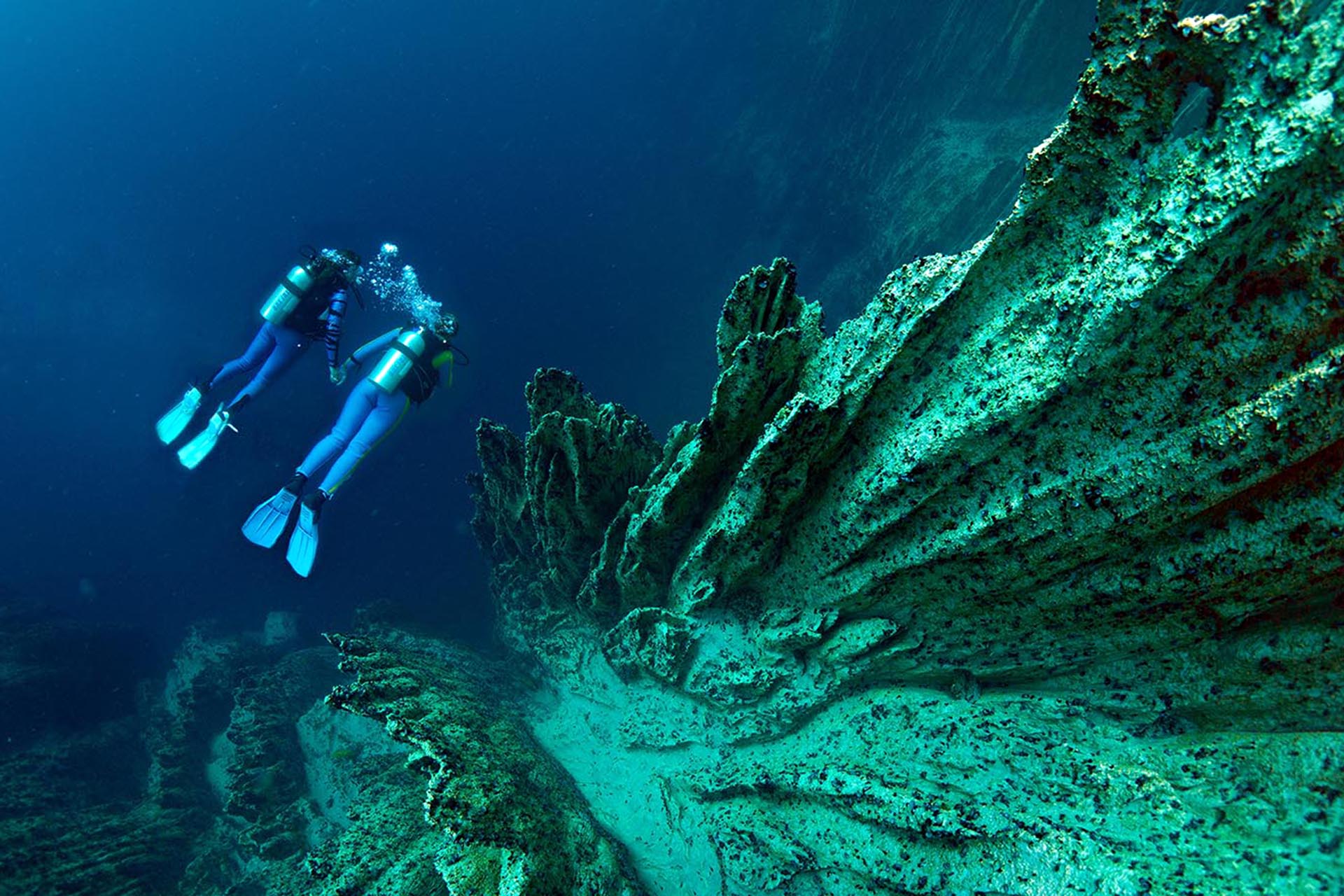 Two divers holding hands underwater and swimming upward next to sharp rock formations.