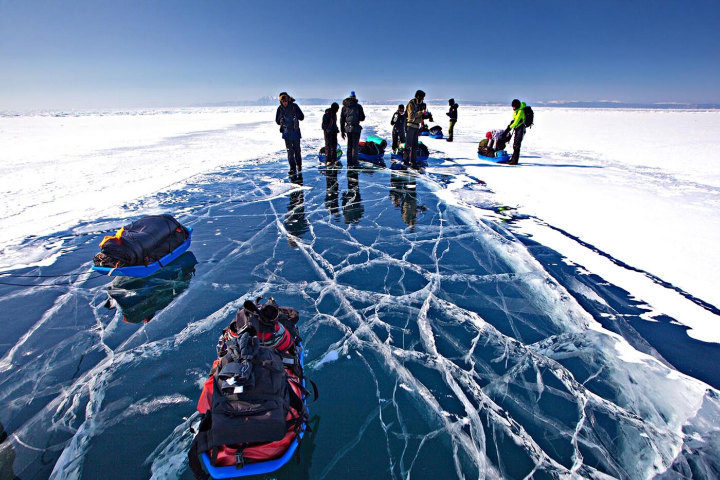 A group of people standing on the cracked ice of a frozen-over lake. They're dragging their packs on sleds.