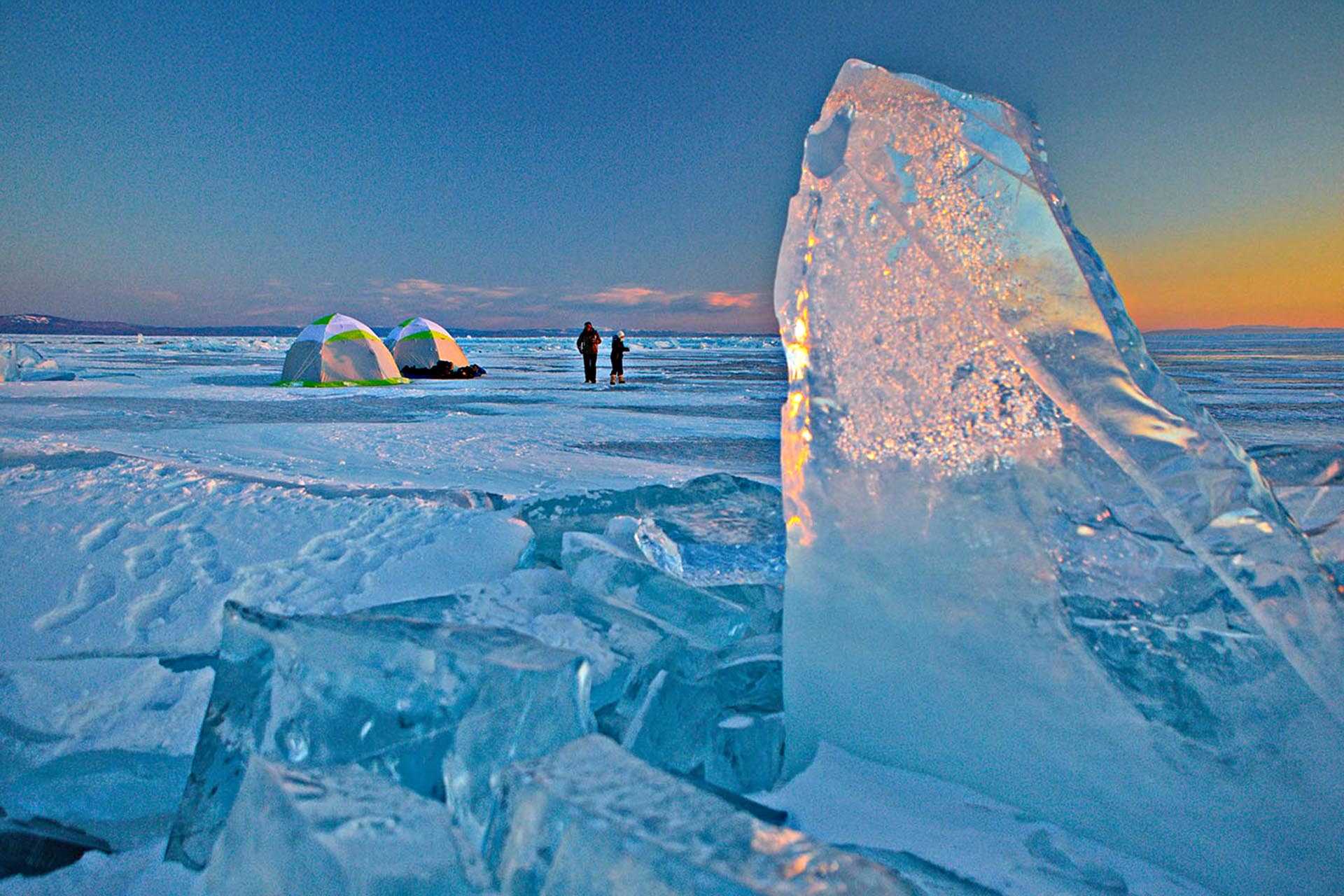 Large formations of ice that reflect the sun to give off an orange hue. In the background, two people and two tents.