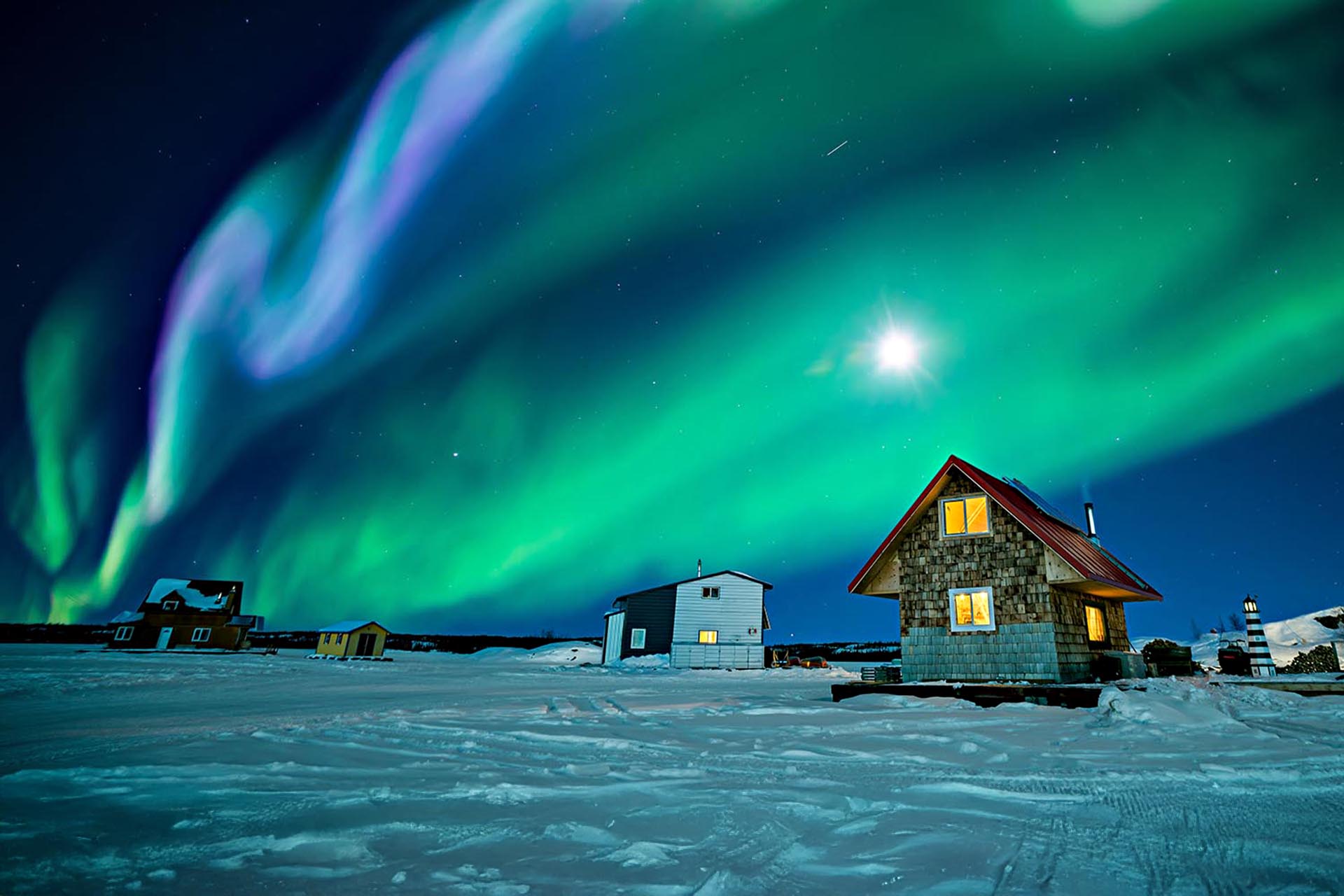 The Aurora Borealis turns the sky green over a group of small houses in the snow.