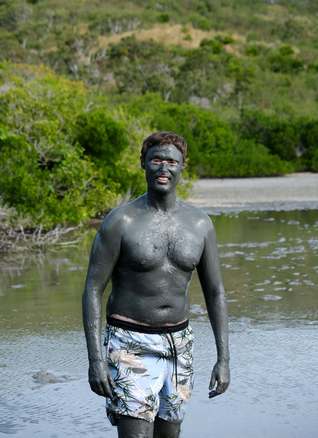 A man standing in the water with grey mud all over him.