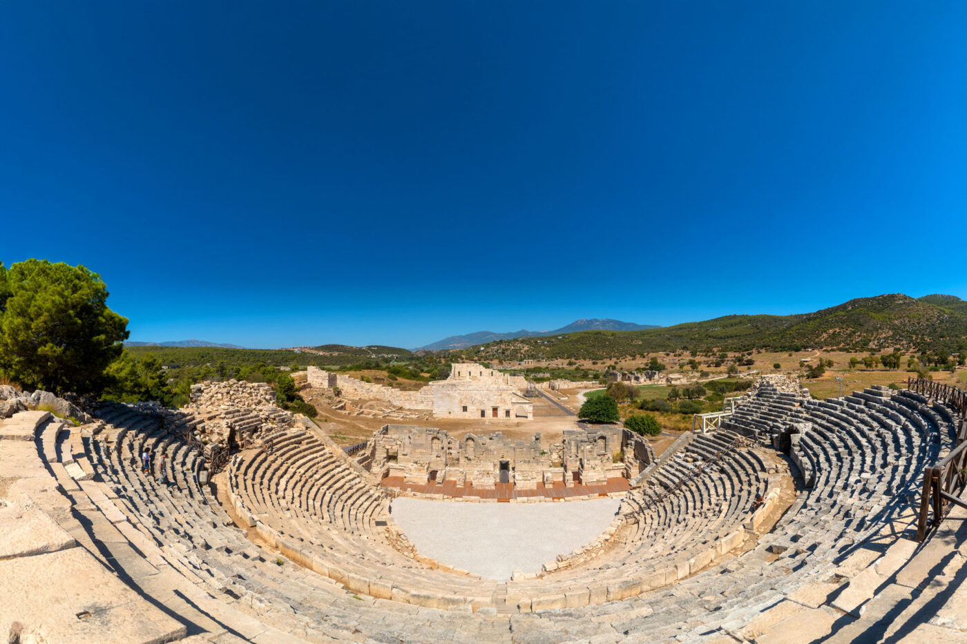 Ruins of a theatre, with seats slanted down to the stage in the middle. In the background, big hills.